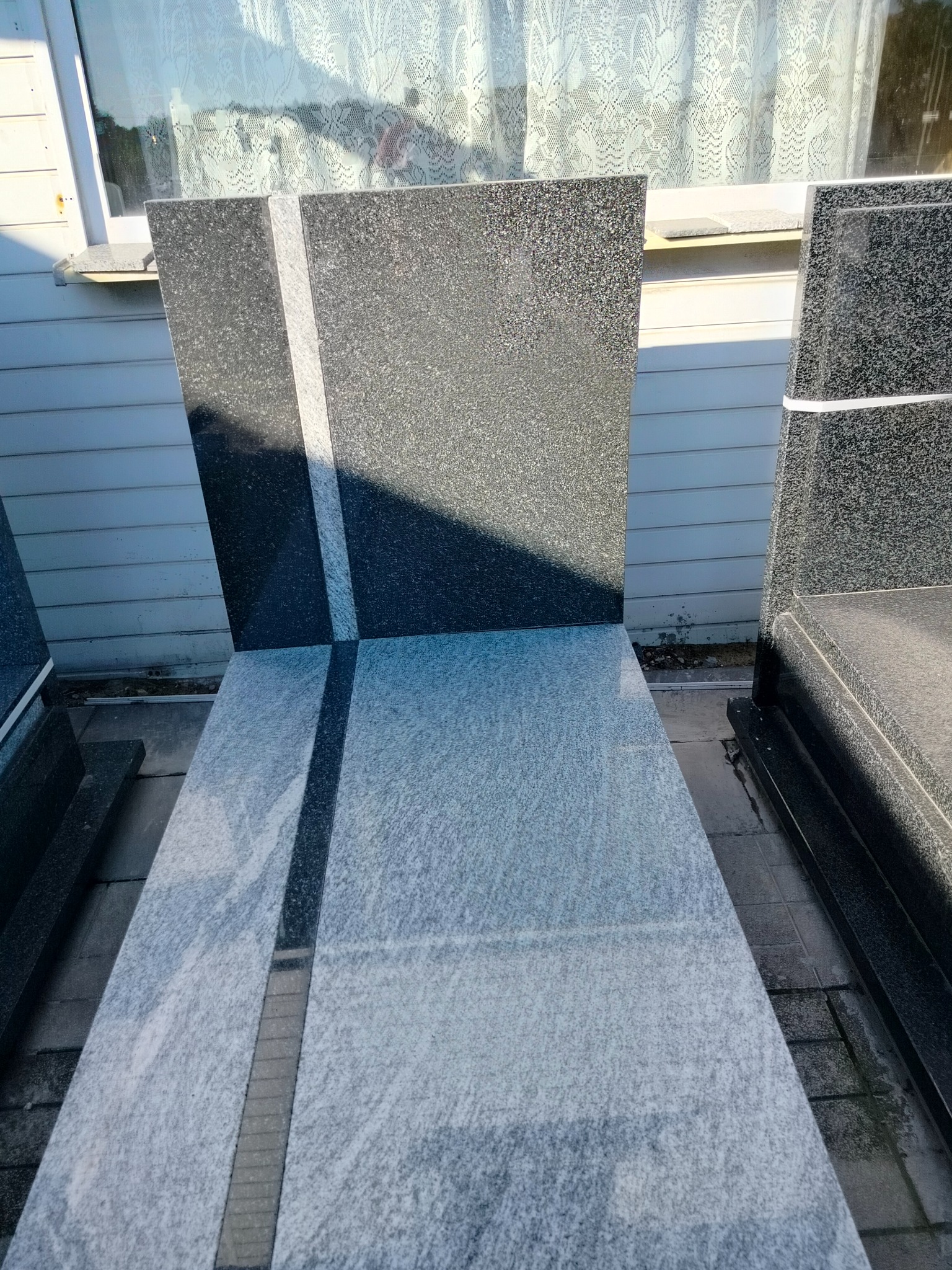 Czarno-szary nagrobek z granitu z jasnym pasem w kształcie krzyża, ustawiony na zewnątrz, w tle biały budynek z oknem.