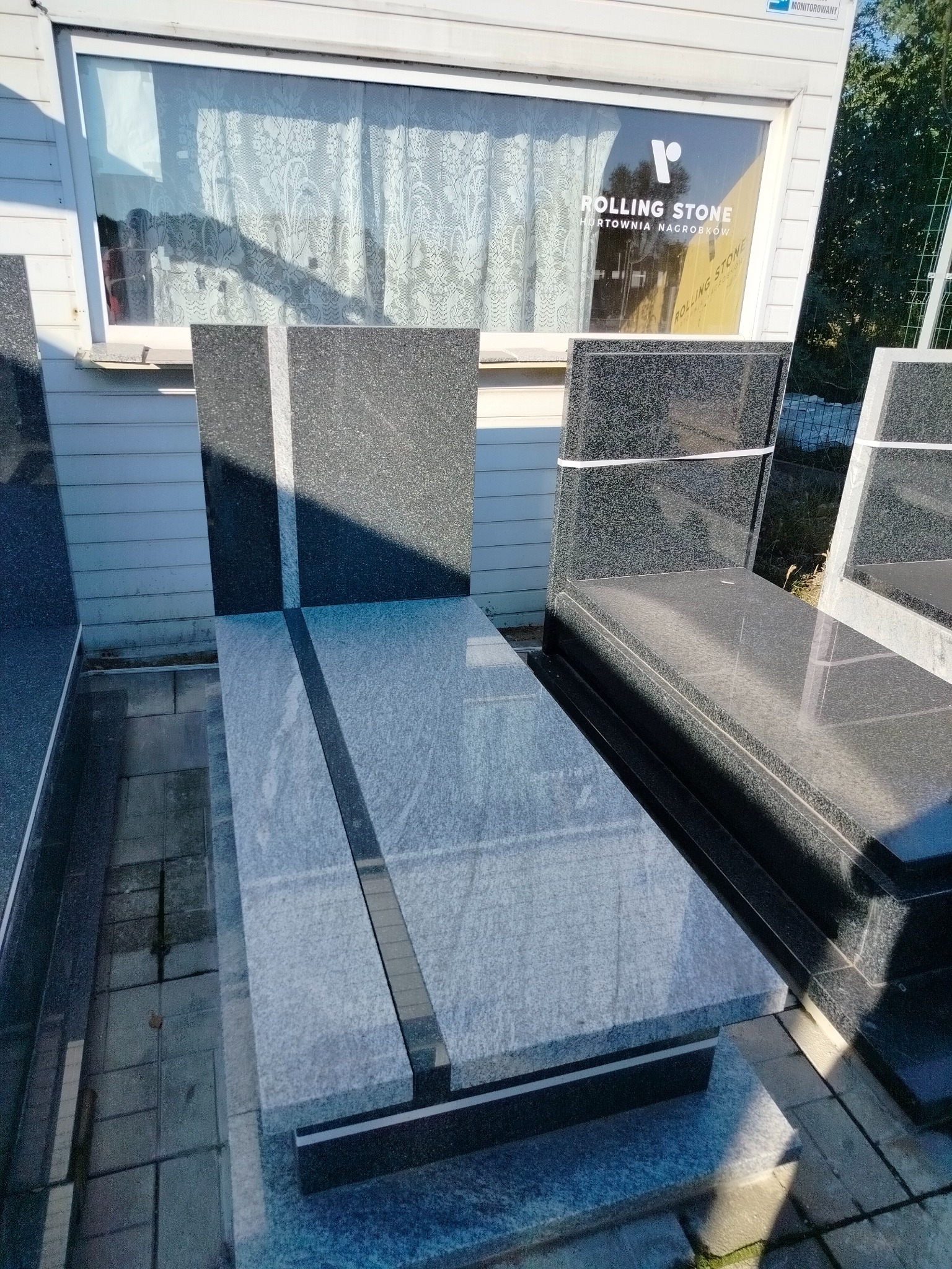 Nowoczesny, szary nagrobek z czarnymi elementami, widoczny z góry, na tle okna z logo firmy kamieniarskiej. Prosta forma, gładkie powierzchnie kamienne.