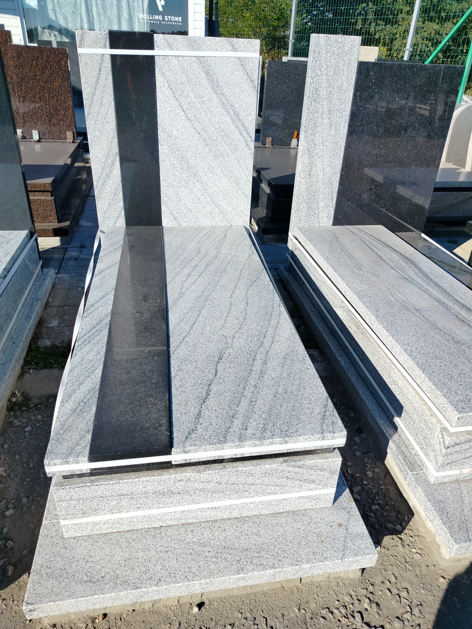 Jasnoszare, granitowe nagrobki z czarnymi elementami, ustawione warstwowo na zewnątrz, w tle widoczne inne nagrobki i zieleń. Ekspozycja wyrobów kamieniarskich.