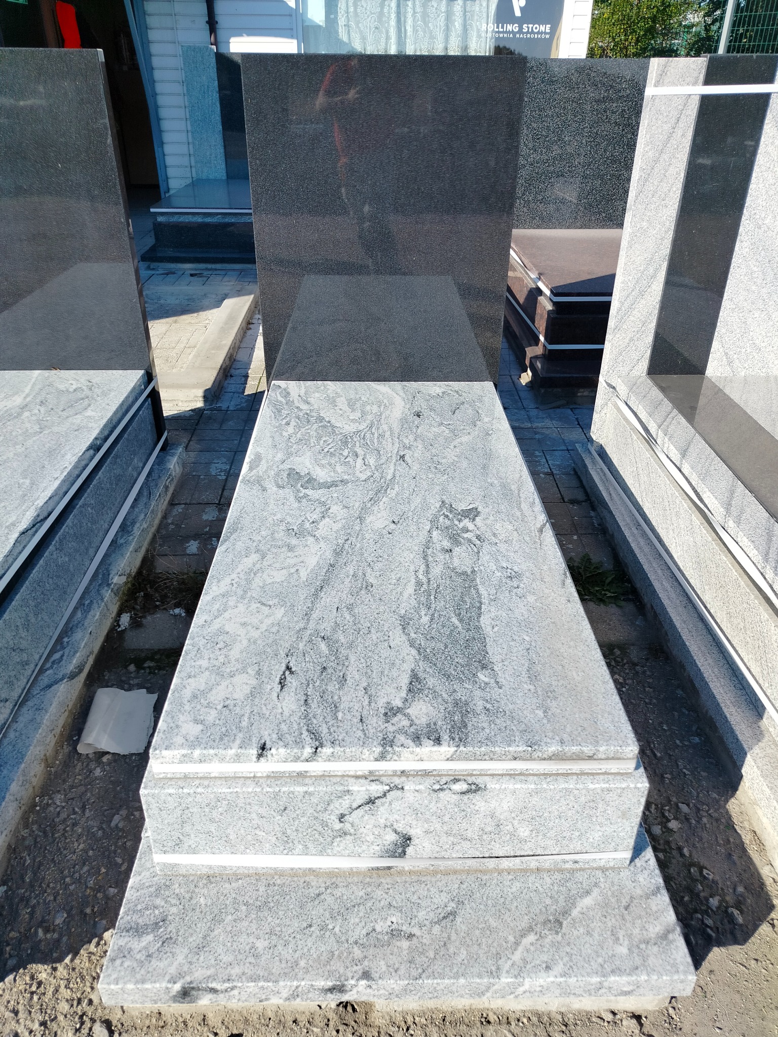 Jasnoszary nagrobek z granitu o unikalnym wzorze, ustawiony na zewnątrz, widoczny z góry. Obok inne elementy nagrobkowe i logo 'Rolling Stone'.