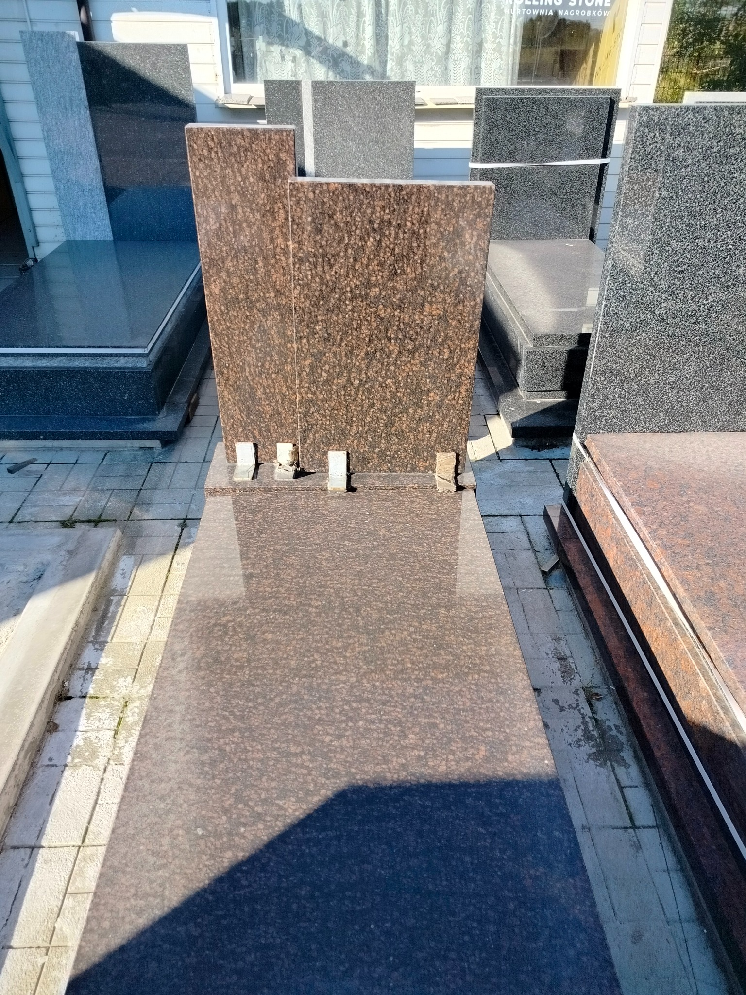 Brązowy nagrobek z granitu w ekspozycji zewnętrznej, widoczne inne nagrobki z różnych kamieni i faktur. Słoneczny dzień.