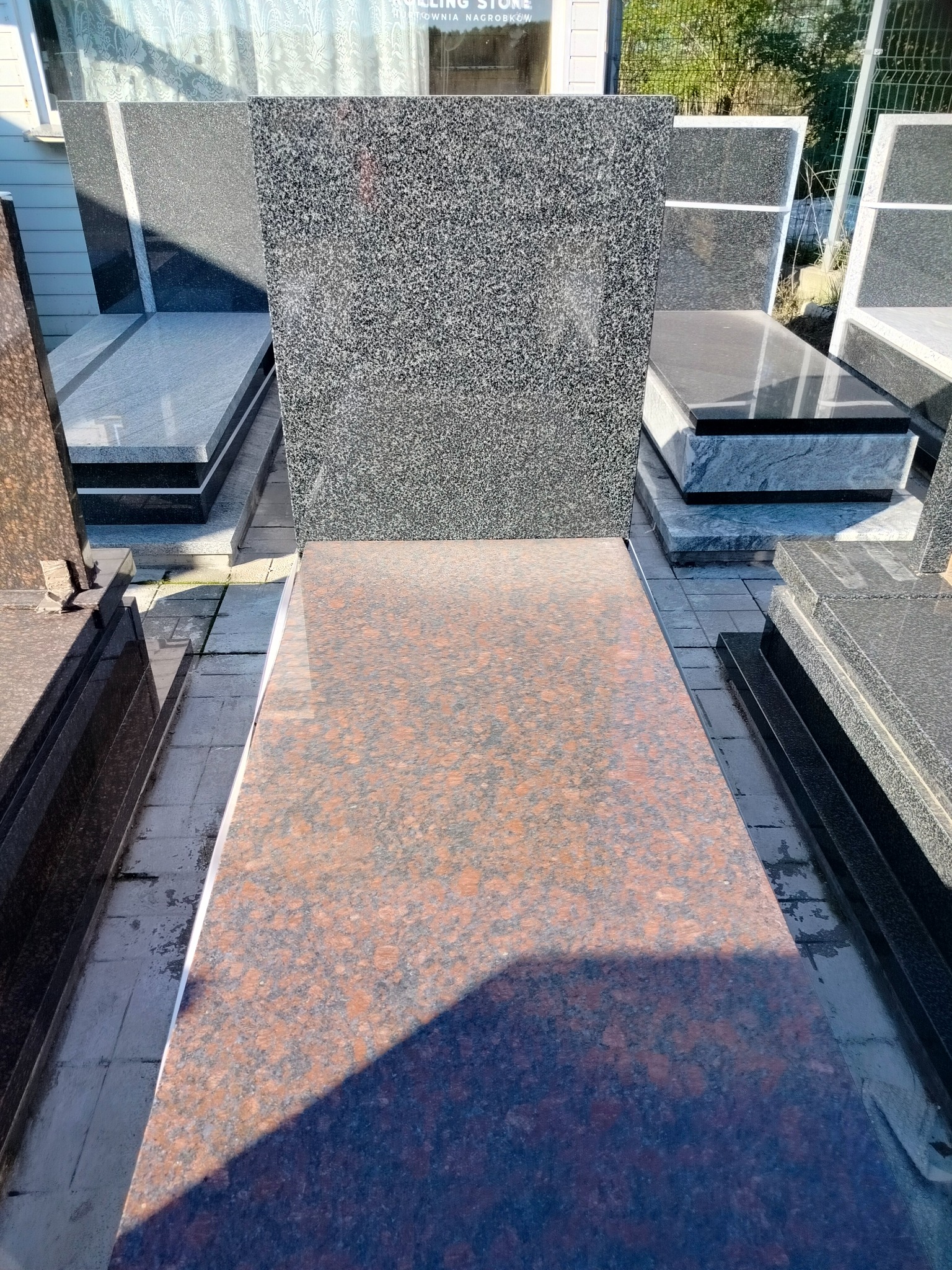 Wystawa nagrobków z granitu o różnych kolorach i fakturach, z przewagą brązowego i szarego, ustawionych na zewnątrz budynku z widocznym napisem 'ROLLING STONE'.