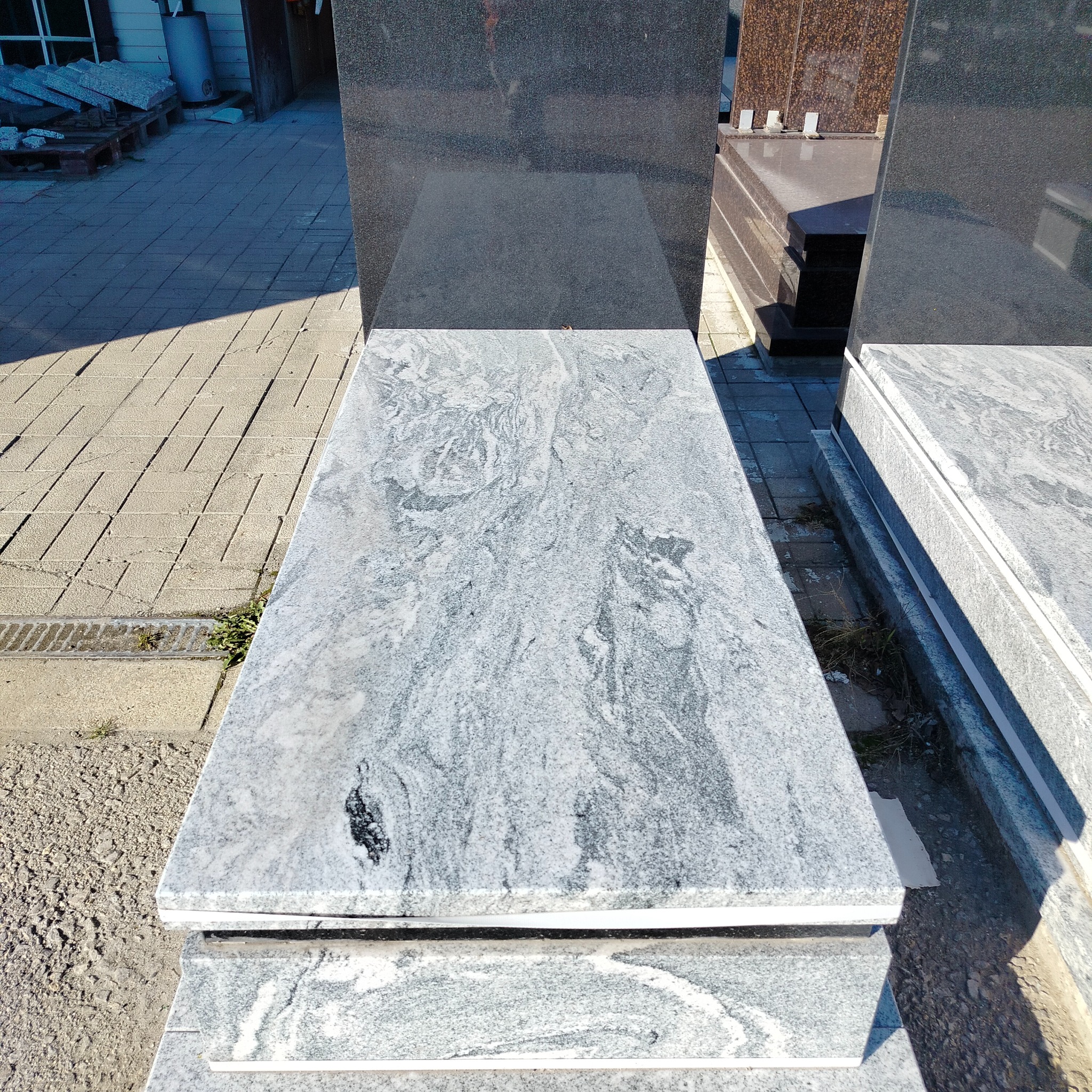 Jasnoszary nagrobek z unikalnym wzorem kamienia, widoczny na zewnątrz w słoneczny dzień. Widoczne inne nagrobki w tle.