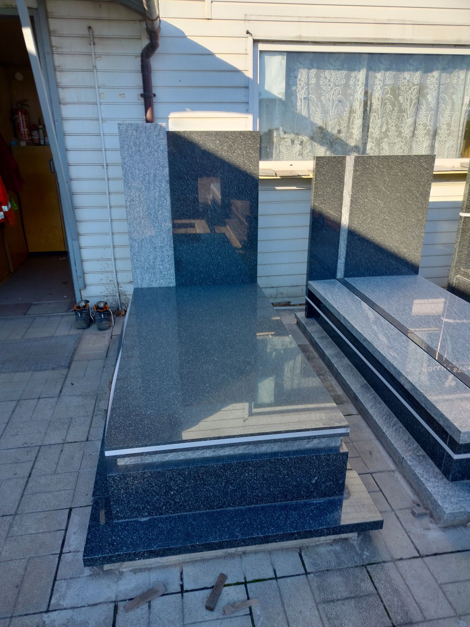 Gotowe nagrobki z ciemnego i szarego kamienia na zewnątrz, w tle budynek z białym sidingiem i oknem z koronkową firanką, buty robocze przy jednym z nagrobków.