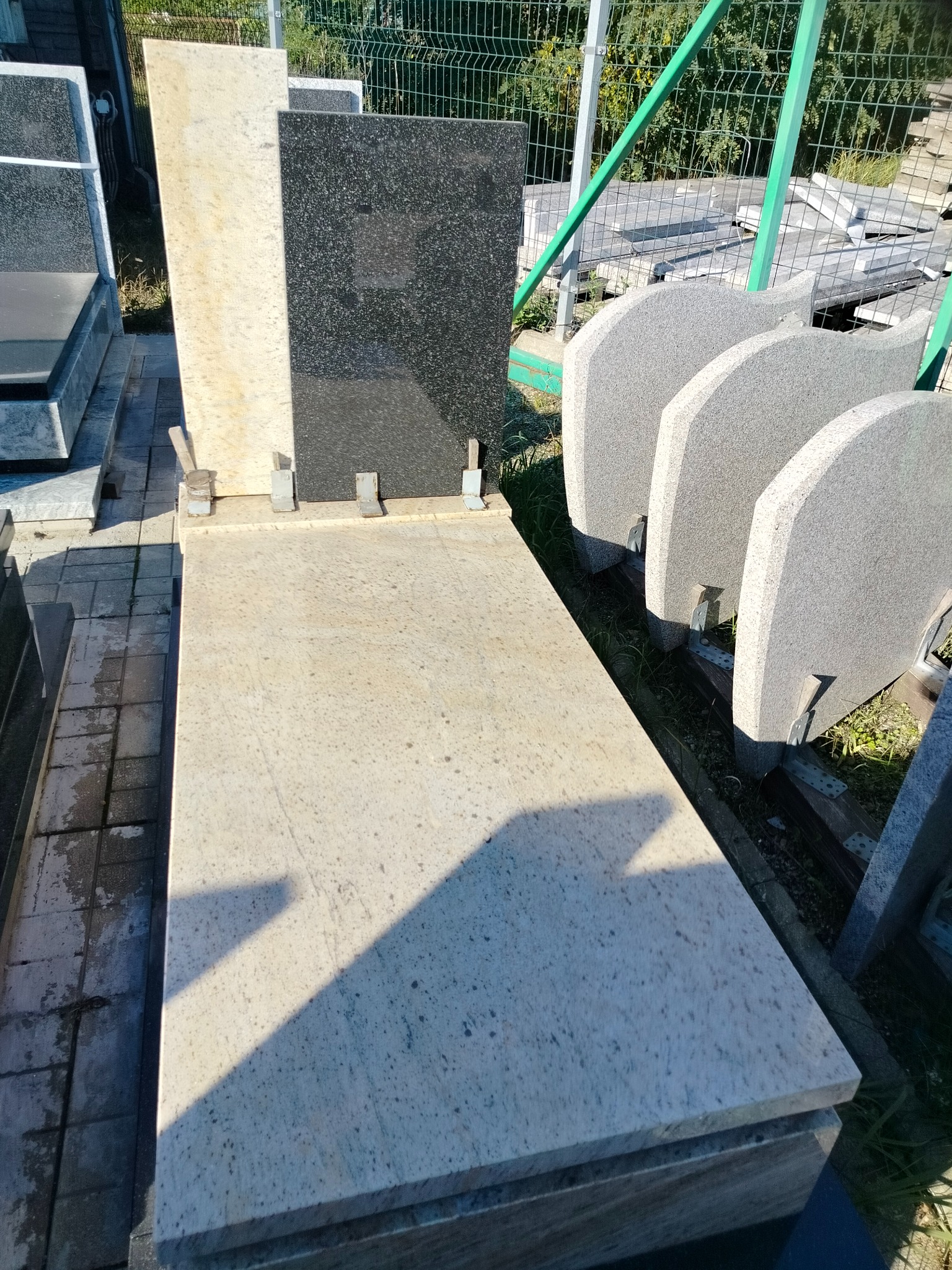 Jasny, gotowy nagrobek z granitu w słoneczny dzień, z czarną tablicą i przygotowanymi elementami nagrobkowymi w tle. Widoczne mocowania i faktura kamienia.