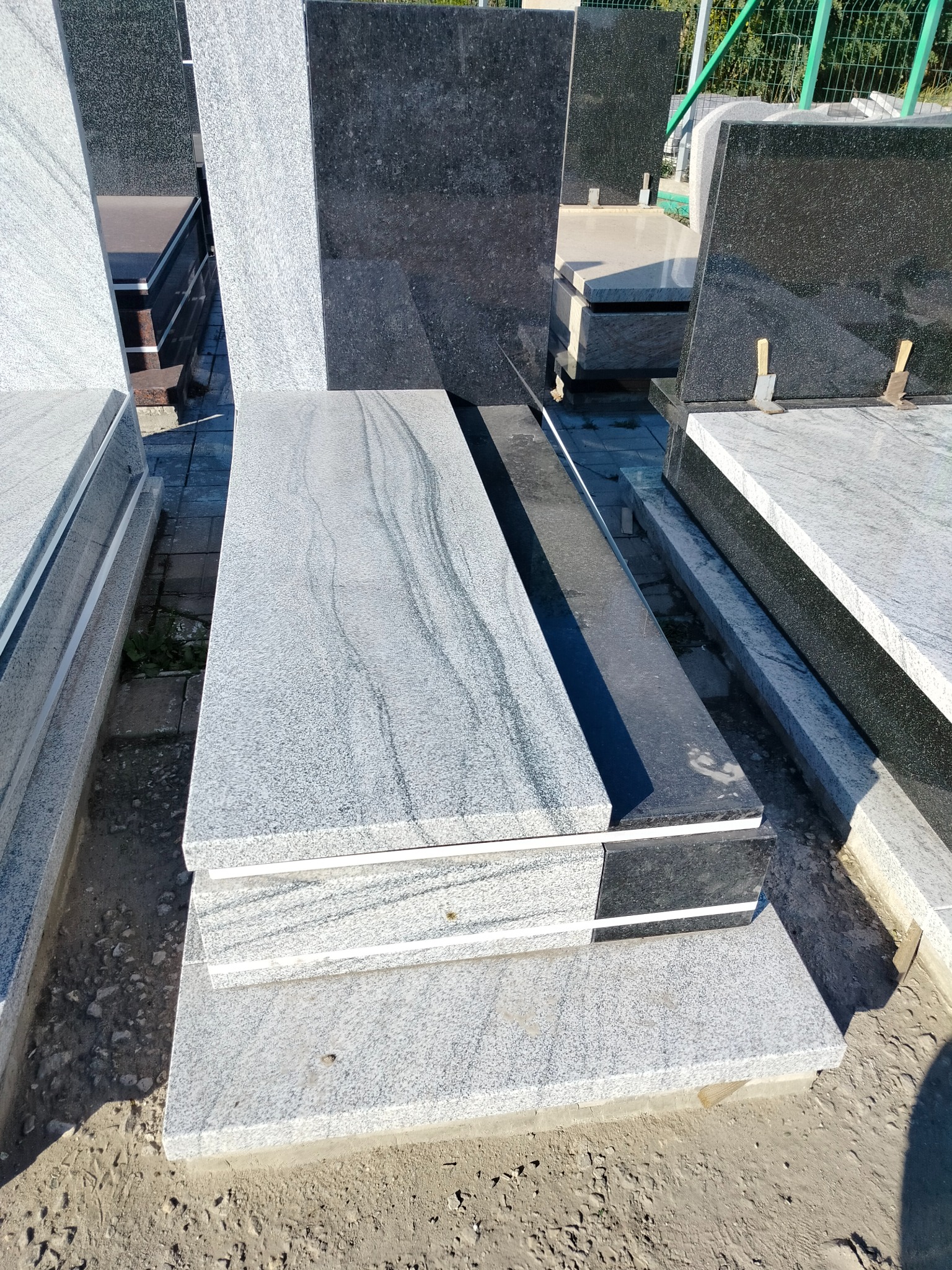 Jasnoszary nagrobek z granitu o wyraźnym wzorze, ustawiony na placu kamieniarskim, obok ciemniejszych, wypolerowanych płyt. Widoczny fragment zieleni w tle.