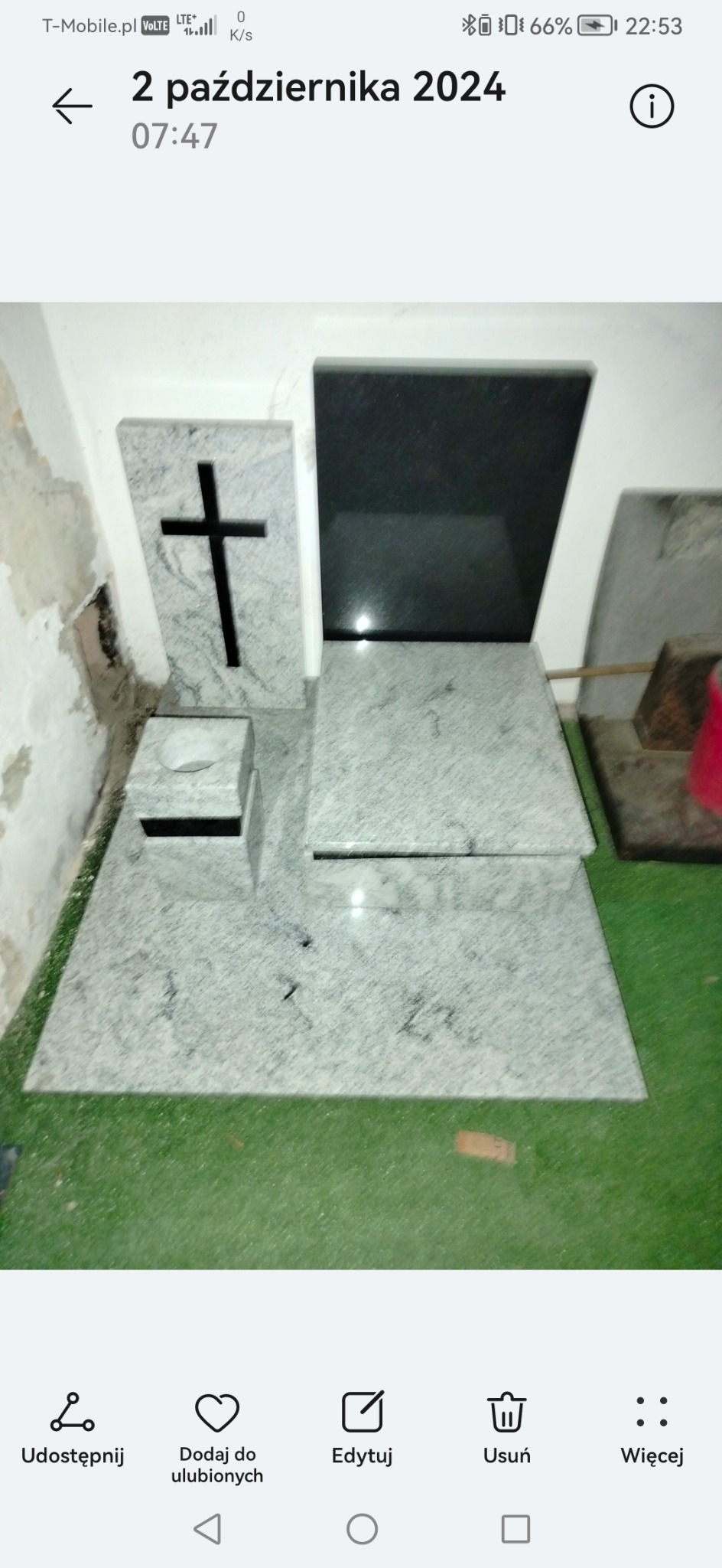 Jasnoszary nagrobek z krzyżem i czarną tablicą, ustawiony na zielonej powierzchni. Widoczne niedoskonałości ściany w tle. Ujęcie z bliska, prezentujące detale kamieni.