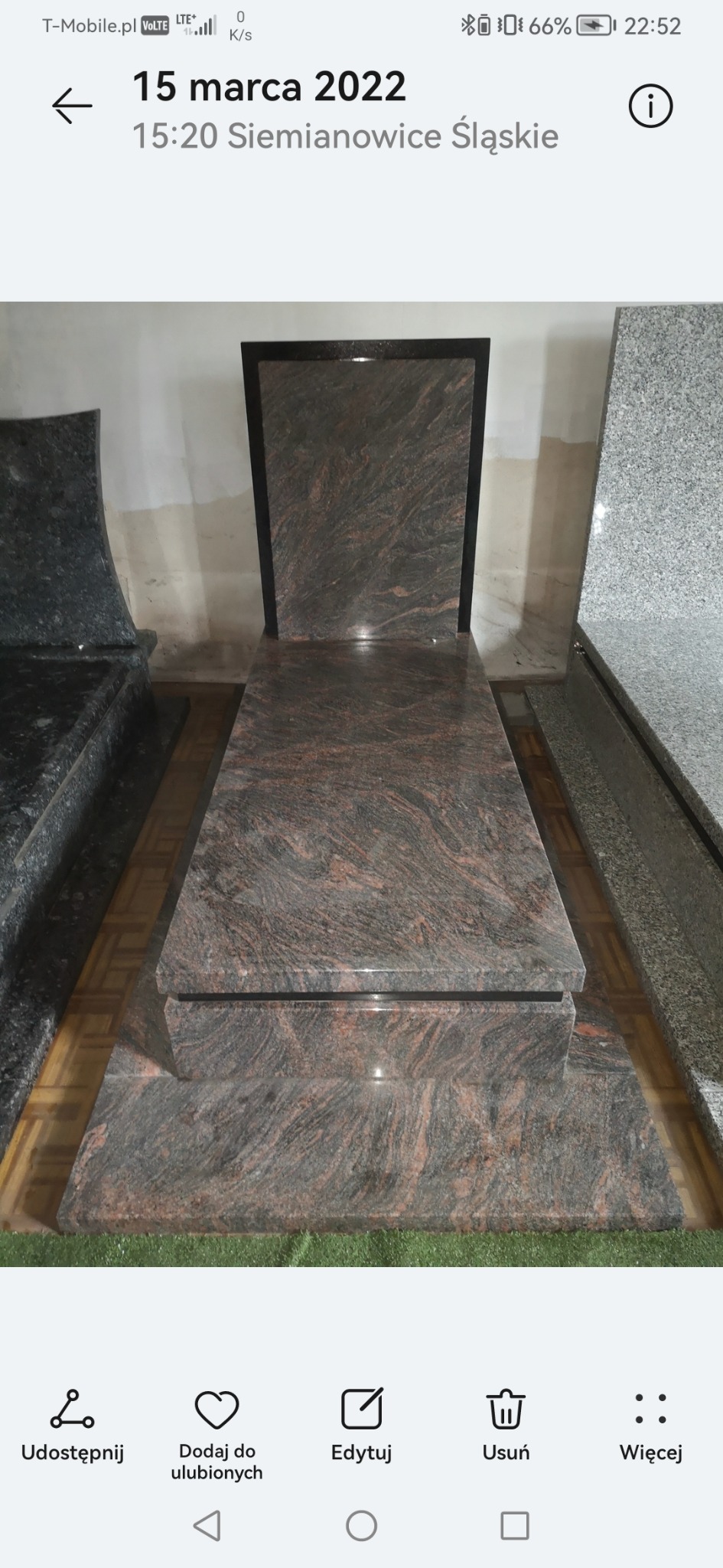 Granitowy nagrobek z ciemnym obramowaniem, prezentowany w zakładzie kamieniarskim. Widoczne inne nagrobki w tle, podłoga wyłożona panelami.