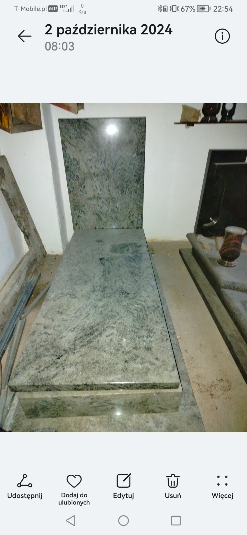 Jasnoszary nagrobek z granitu o wyraźnym wzorze, ustawiony w pomieszczeniu. Widoczne elementy innego nagrobka w tle.