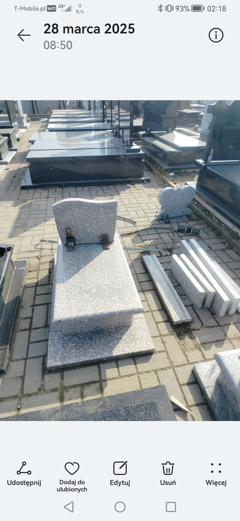 Jasnoszary, granitowy nagrobek z krzyżem i dodatkowymi elementami, ustawiony na placu z kostki brukowej. W tle inne nagrobki i elementy kamienne.