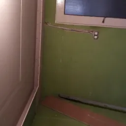 Wnętrze pokoju w trakcie remontu: ściana z zamontowaną płytą kartonowo-gipsową, widoczne otwory po wkrętach, surowa zielona ściana z wystającą instalacją elektryczną i oknem, poziomica i fragment...