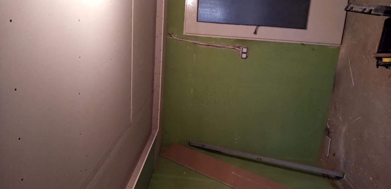 Wnętrze pokoju w trakcie remontu: ściana z zamontowaną płytą kartonowo-gipsową, widoczne otwory po wkrętach, surowa zielona ściana z wystającą instalacją elektryczną i oknem, poziomica i fragment...