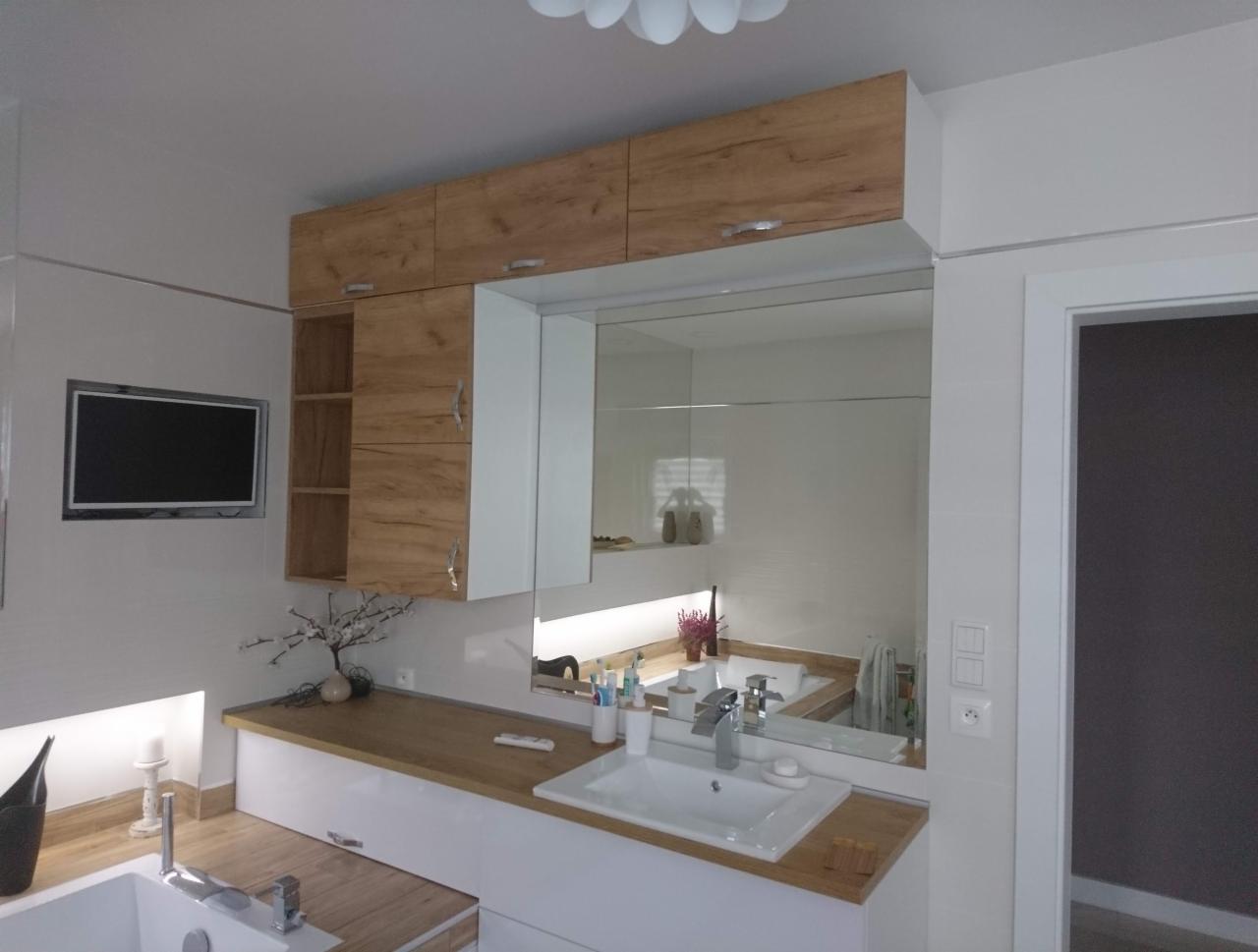 Nowoczesna łazienka z drewnianymi szafkami nad umywalką, dużym lustrem, wbudowanym telewizorem w ścianie i dekoracyjnym drzewkiem w wazonie.