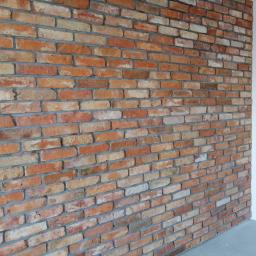 Lemkaz - Ściana wykończona cegłą klinkierową w odcieniach czerwieni, beżu i bieli, z szarymi spoinami, przylegająca do białej ściany i szarej posadzki.