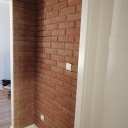 Lemkaz - Ściana z cegły klinkierowej w korytarzu w Czernicy, z widocznymi białymi włącznikami światła i białymi listwami przypodłogowymi, częściowo widoczne drzwi z białymi ościeżnicami.