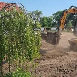 Żółta koparka Mecolac w trakcie prac ziemnych na działce, widoczny pracownik z łopatą oraz przygotowany teren pod budowę domu z czerwoną dachówką.