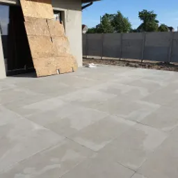 Jasnoszary taras betonowy ułożony z dużych płyt, z widocznym fragmentem budynku w stanie surowym i tymczasowymi zabezpieczeniami okien z płyt OSB, ogrodzenie panelowe w tle.