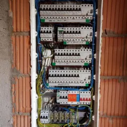 Rozdzielnica elektryczna z licznymi bezpiecznikami i przewodami, zamontowana w ścianie z cegły dziurawki, widoczne pianka montażowa i przewody w peszlu.