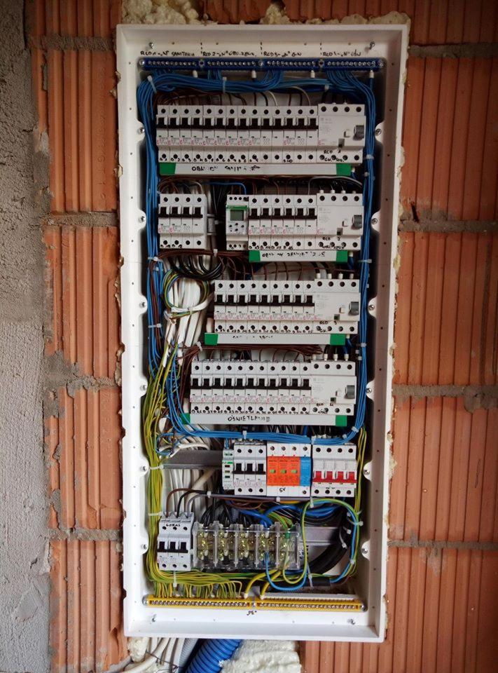 Rozdzielnica elektryczna z licznymi bezpiecznikami i przewodami, zamontowana w ścianie z cegły dziurawki, widoczne pianka montażowa i przewody w peszlu.