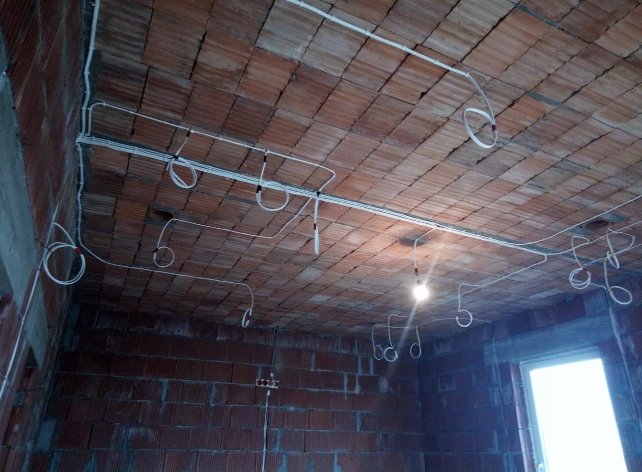 Instalacja elektryczna w surowym wnętrzu: białe kable prowadzone po ceglanym suficie, tymczasowe oświetlenie z żarówki, widoczne puszki elektryczne na ścianie, okno w stanie surowym.