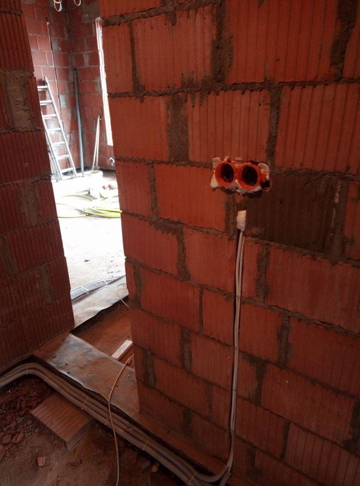 Surowa ściana z czerwonej cegły z zamontowanymi puszkami elektrycznymi i prowadzoną instalacją kablową, widoczne przewody w peszlu na podłodze, w tle drabina w pomieszczeniu w stanie deweloperskim.