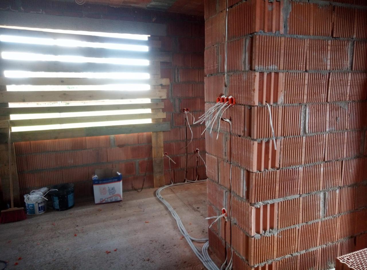 Surowa instalacja elektryczna w budynku z czerwonej cegły, widoczne kable wystające z puszek montażowych, okno zabite deskami, podłoga w stanie surowym.