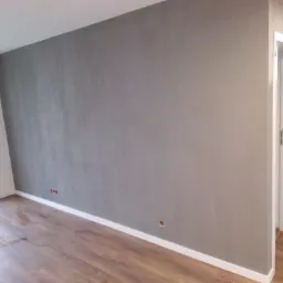 Pokój po malowaniu z szarą ścianą o fakturze betonu, widoczne gniazdka elektryczne i drewniana podłoga, białe listwy przypodłogowe i otwarte białe drzwi do łazienki z czarno-białą mozaiką.