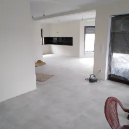 Jasne, wykafelkowane wnętrze w trakcie prac wykończeniowych, z widoczną maszyną do polerowania pod oknem zabezpieczonym folią i prowizorycznym zabezpieczeniem podłogi z kartonu.