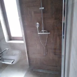 Kabina prysznicowa z brązowymi płytkami ułożonymi w jodełkę, widoczny odpływ liniowy, bateria prysznicowa z deszczownicą i słuchawką oraz fragment toalety za szybą kabiny.