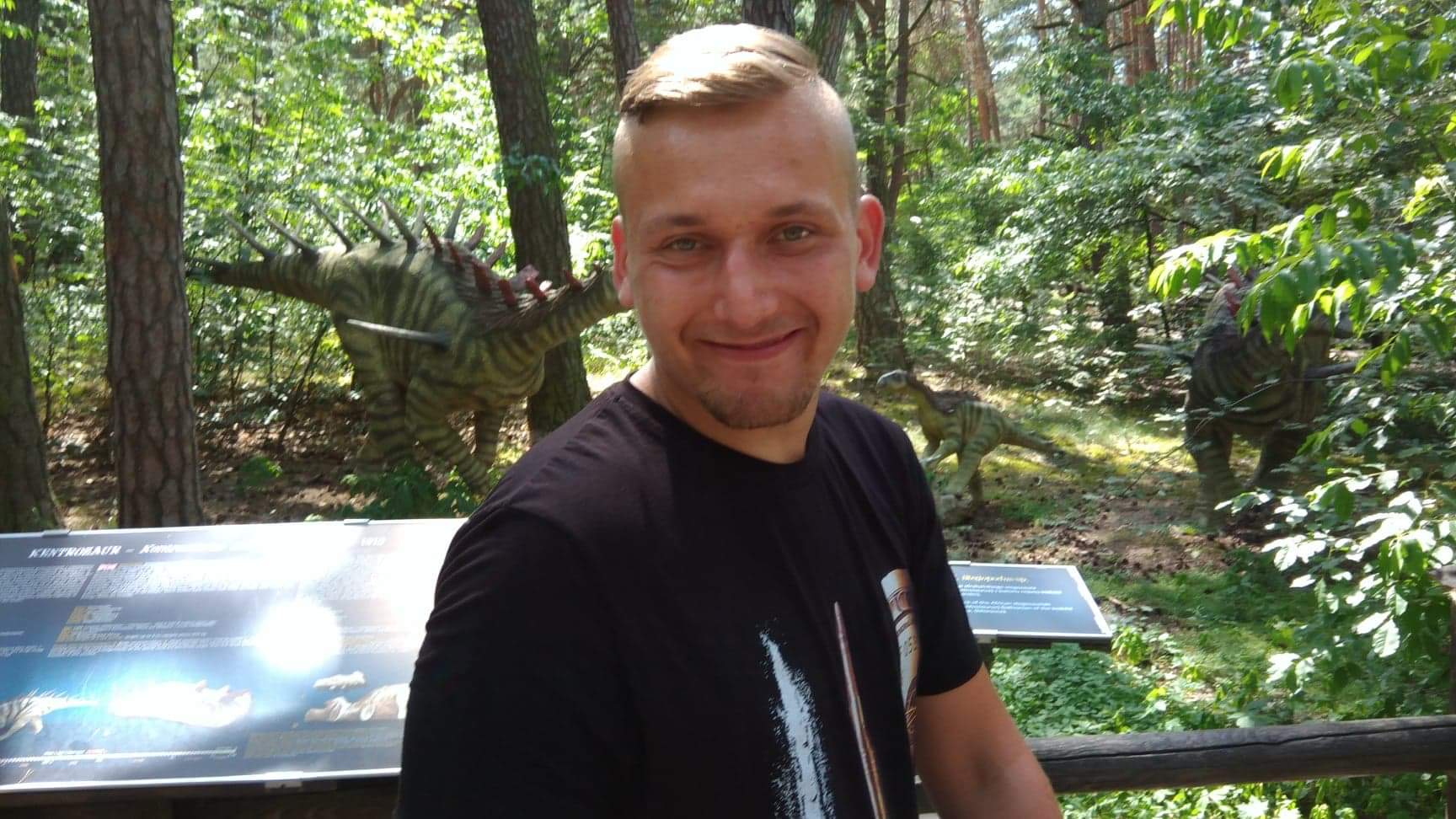 Uśmiechnięty mężczyzna na tle parku dinozaurów, z widocznymi modelami Kentrozaura i innych. Tablica informacyjna w kadrze, drzewa i zieleń w tle.