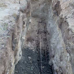 EURO INVESTIA SPÓŁKA Z OGRANICZONĄ ODPOWIEDZIALNOŚCIĄ - Wykop pod fundament ogrodzenia z umieszczonym zbrojeniem w postaci metalowej klatki, częściowo wypełniony gruzem i ziemią.