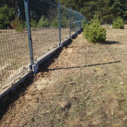 EURO INVESTIA SPÓŁKA Z OGRANICZONĄ ODPOWIEDZIALNOŚCIĄ - Długi fragment świeżo zamontowanego ogrodzenia z siatki panelowej na betonowej podmurówce, widoczne ślady prac ziemnych i młode drzewka iglaste w tle.