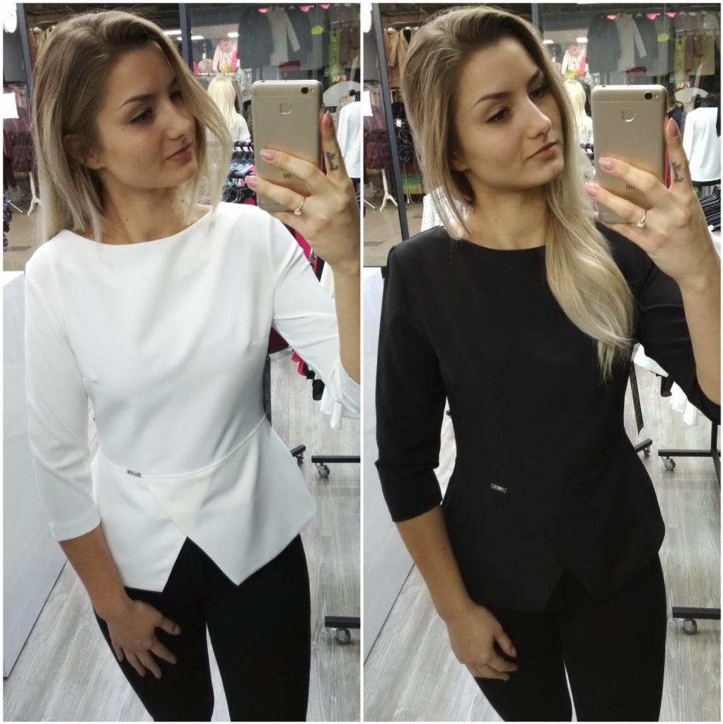 Selfie kobiety prezentującej białą i czarną bluzkę z asymetrycznym dołem na tle sklepowego wieszaka z ubraniami, widoczny tatuaż na palcu.