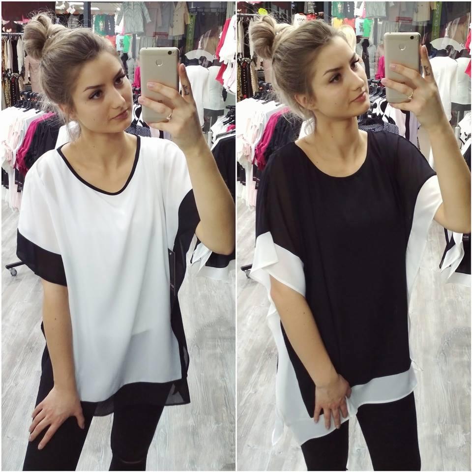 Kobieta robi selfie w lustrze, prezentując dwa warianty tej samej tuniki: białą z czarnymi wstawkami i czarną z białymi wstawkami, na tle wieszaków z odzieżą w sklepie.