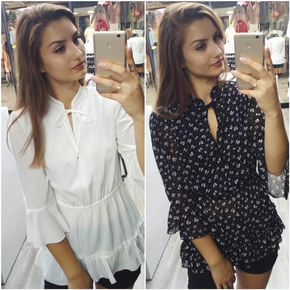 Selfie kobiety prezentującej dwie bluzki z falbanami: białą z wiązaniem i czarną w drobny wzór, z widoczną ekspozycją sklepową w tle.