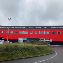 Czerwono-czarny budynek Centrum Budownictwa Jerozolimskie z szyldami reklamowymi, widok z ulicy w pochmurny dzień. Zaparkowane samochody i zielony teren przed budynkiem.