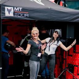 Everyday Events Filip Tucki - Dwie uśmiechnięte kobiety w Zawodnem prezentują stoisko MT Tour na tle autobusu. Stoisko z czarnym zadaszeniem i logo.