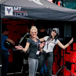 Dwie uśmiechnięte kobiety w Zawodnem prezentują stoisko MT Tour na tle autobusu. Stoisko z czarnym zadaszeniem i logo.