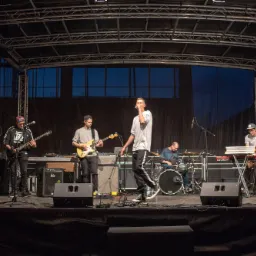 Zespół muzyczny gra na scenie pod zadaszeniem, wokalista w białej koszulce śpiewa do mikrofonu, gitarzysta, basista, klawiszowiec i perkusista grają na instrumentach, widoczne głośniki, kable...