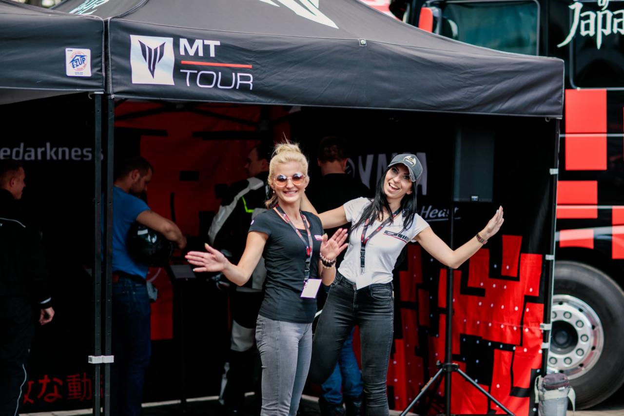 Dwie uśmiechnięte kobiety w Zawodnem prezentują stoisko MT Tour na tle autobusu. Stoisko z czarnym zadaszeniem i logo.