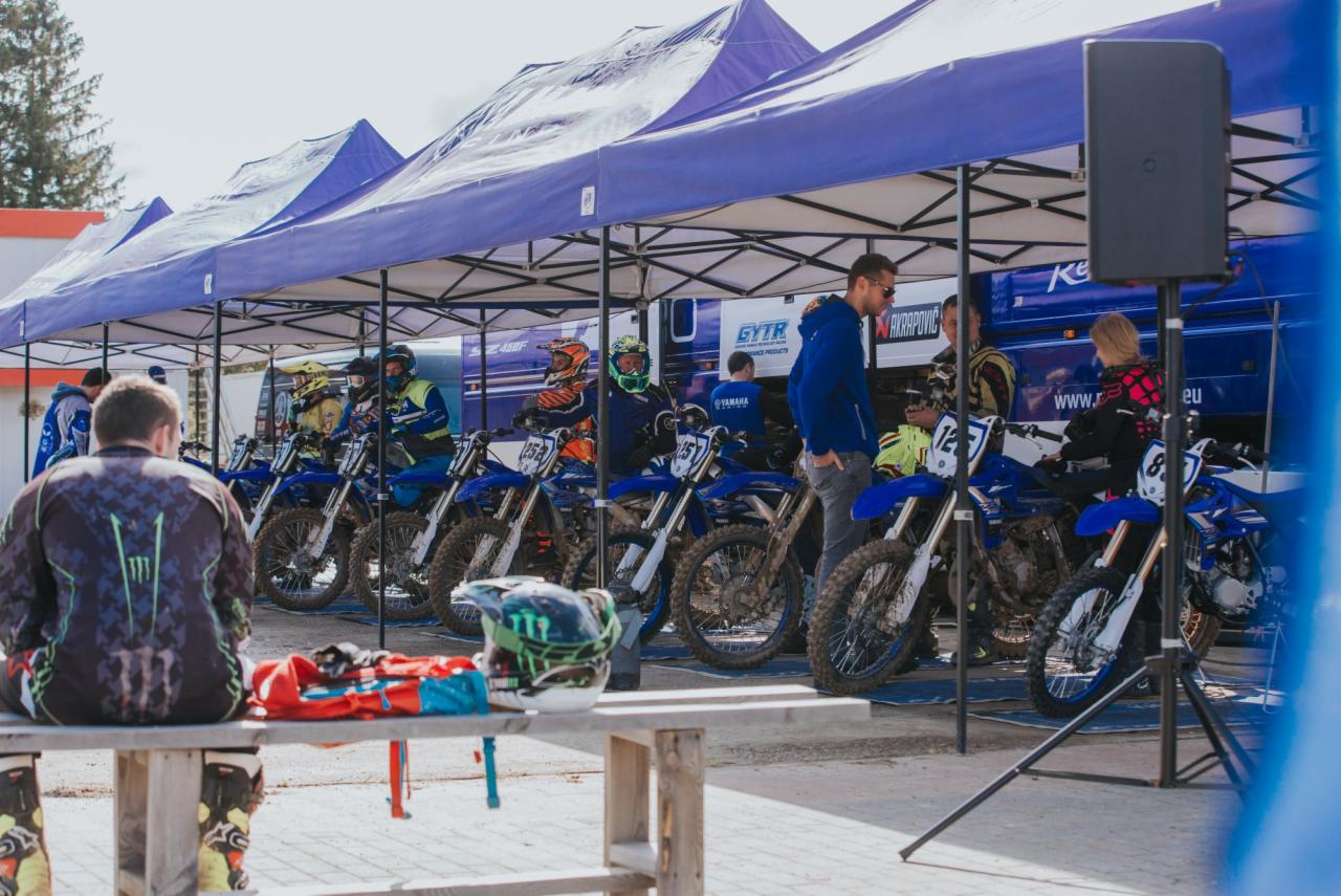 Rząd motocykli motocrossowych Yamaha zaparkowanych pod namiotami, przygotowanych do zawodów, z widocznym sprzętem i zawodnikami w tle.