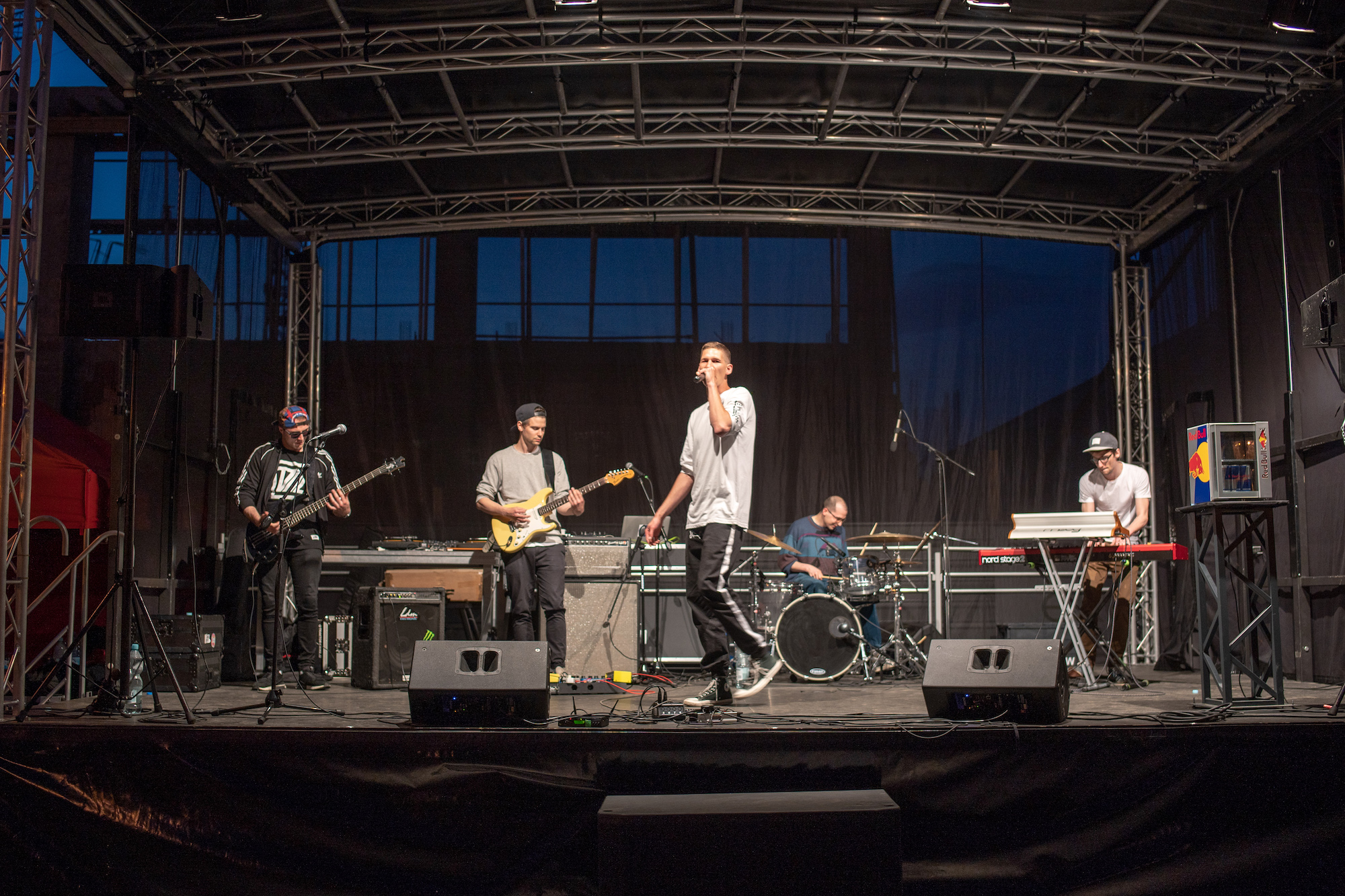 Zespół muzyczny gra na scenie pod zadaszeniem, wokalista w białej koszulce śpiewa do mikrofonu, gitarzysta, basista, klawiszowiec i perkusista grają na instrumentach, widoczne głośniki, kable...