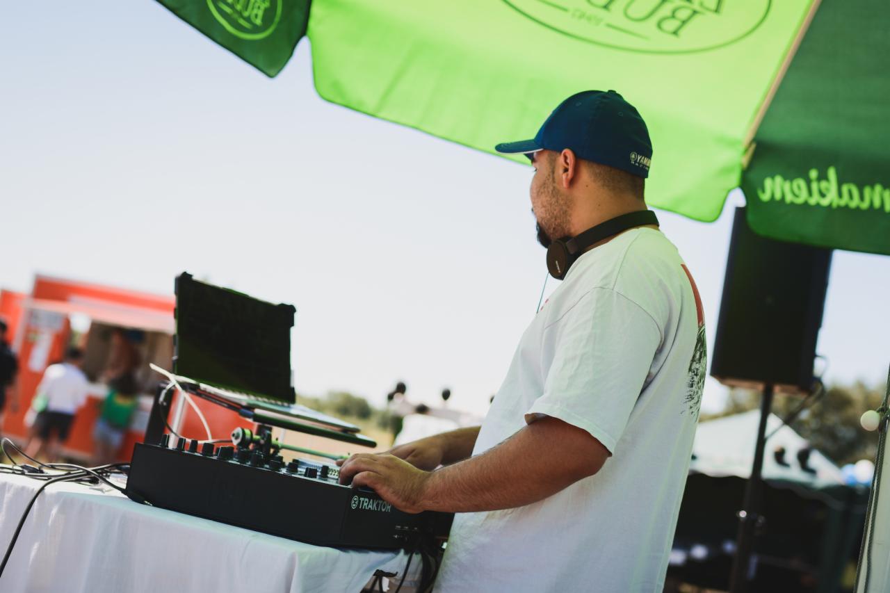 DJ w niebieskiej czapce obsługuje mikser Traktor na zewnątrz, pod zielonym parasolem reklamowym, z widocznym food truckiem w tle.