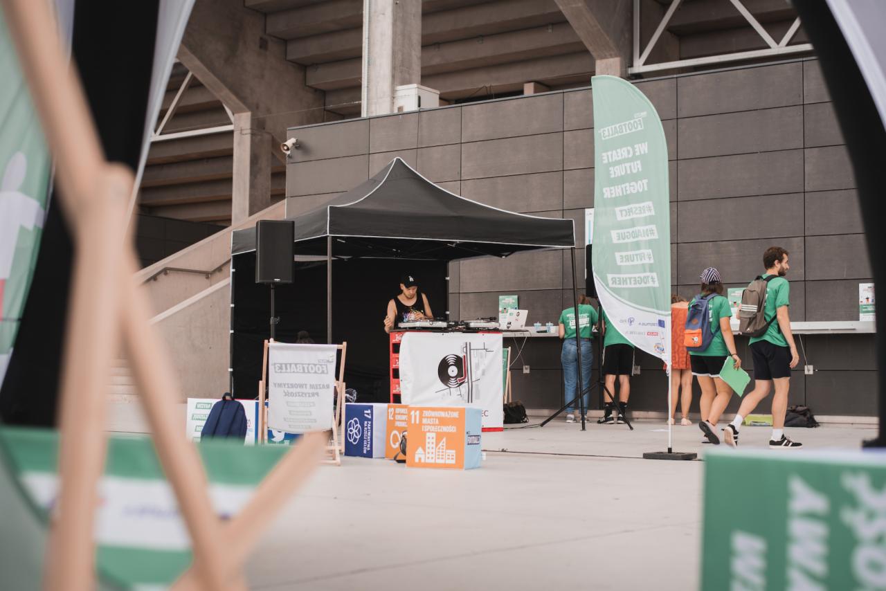 DJ grający muzykę na imprezie plenerowej pod zadaszeniem, widoczny sprzęt nagłośnieniowy, flaga z hasłami eventowymi i przechodzący ludzie w tle.