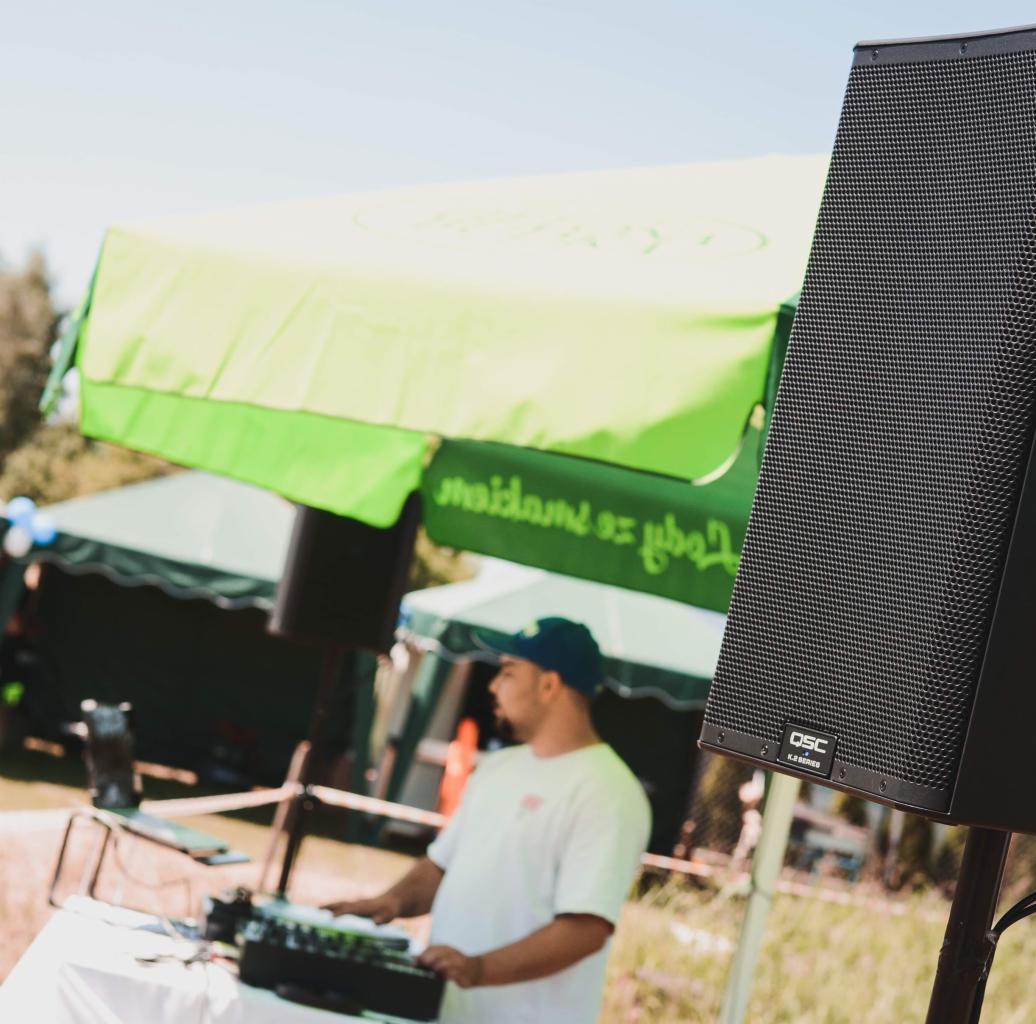 DJ obsługujący sprzęt nagłośnieniowy QSC K.2 Series na zewnątrz, w tle zielony namiot i częściowo widoczna scena.