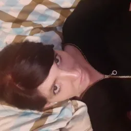 Kobieta leżąca na ł&oacute;żku w czarnej bluzie z suwakiem, patrząca w g&oacute;rę, z widocznymi brązowymi włosami i fragmentem wzorzystej pościeli.