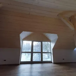Poddasze z drewnianym wykończeniem, widoczne okno z nietypowym kształtem i drewniana belka konstrukcyjna, surowy stan wykończenia.