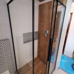 Usługi Budowlano Remontowe Michał Powęska - Nowoczesna kabina prysznicowa typu walk-in z czarnymi profilami, deszczownicą i drewnopodobną okładziną ściany, z widocznym odpływem liniowym w podłodze.