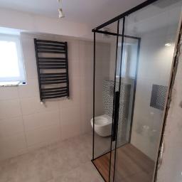 Usługi Budowlano Remontowe Michał Powęska - Nowoczesna łazienka z kabiną prysznicową typu walk-in z czarnymi profilami, grzejnikiem drabinkowym i płytkami imitującymi beton. W tle widoczna toaleta i fragment ściany z dekoracyjnymi płytkami.