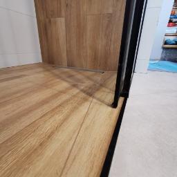 Usługi Budowlano Remontowe Michał Powęska - Fragment kabiny prysznicowej z drewnopodobną okładziną ścienną i podłogową, widoczny odpływ liniowy, czarna rama kabiny.